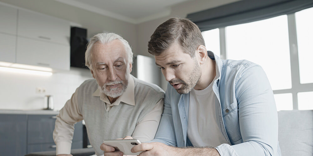 Two men, one older with gray hair and a beard and one younger with brown hair and stubble, sit together indoors on a couch, looking intently at a smartphone in the younger mans hands.