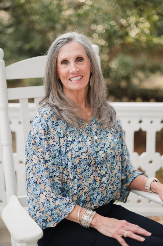 Smiling older woman with long gray hair, wearing a blue floral blouse and silver bracelets, sits on a white rocking chair outdoors with greenery in the background.