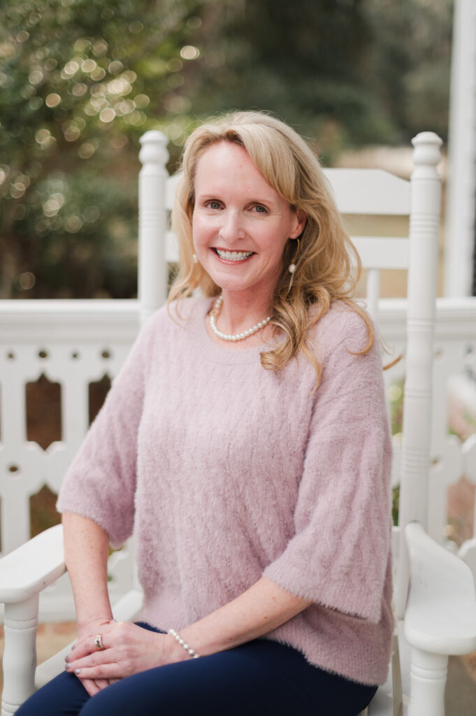 A woman with long blonde hair, wearing a light pink sweater and pearl necklace, sits smiling on a white rocking chair outdoors with greenery in the blurred background.