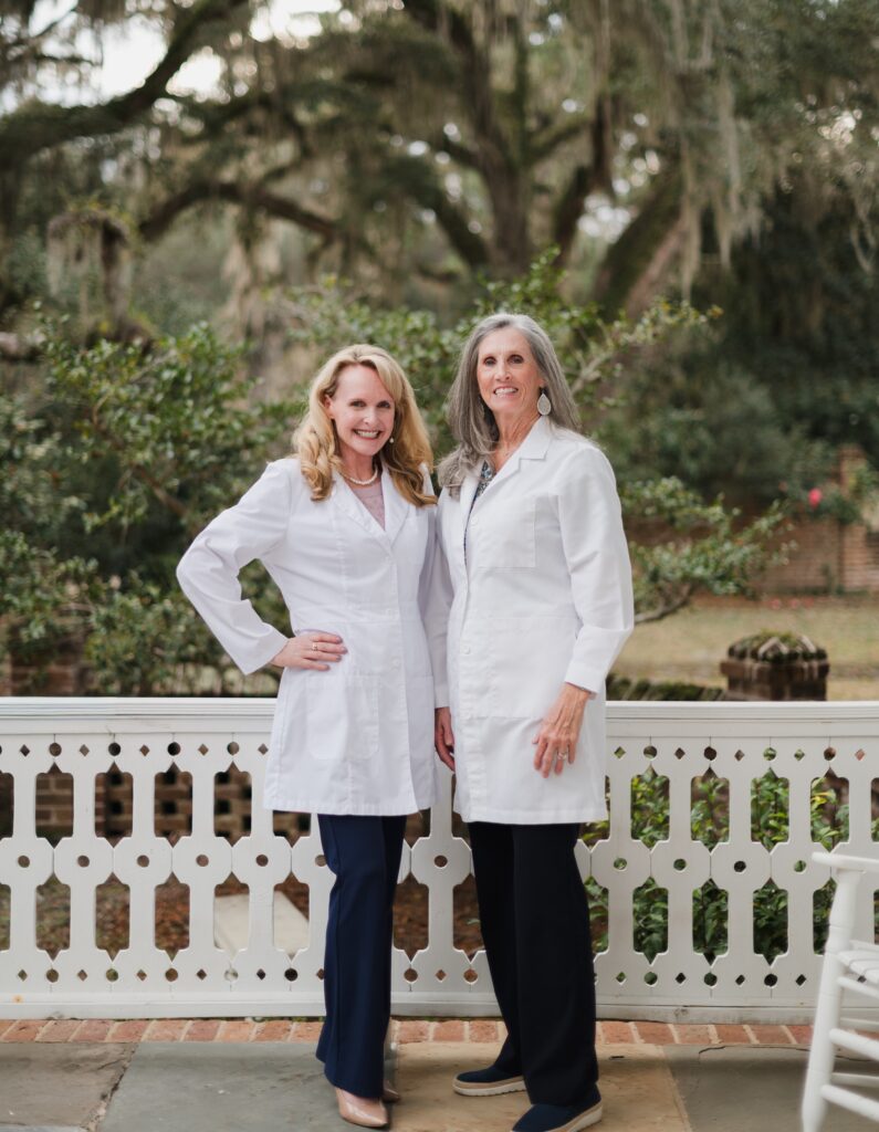 Two women with long hair stand outside on a porch, both smiling and wearing white lab coats and dark pants, ready to support those affected by hearing loss. Lush green trees and a decorative white railing are in the background.