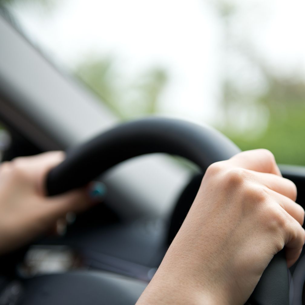 Close-up of a person’s hands gripping a steering wheel inside a car, with a blurred background seen through the windshield.