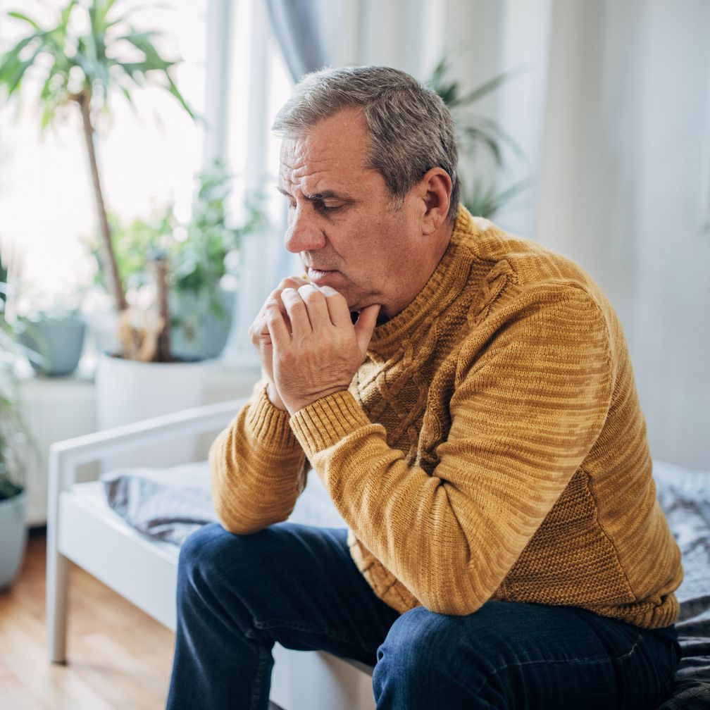 An older man in a yellow sweater sits on a bed with clasped hands, looking thoughtful or worried. There are houseplants and natural light in the background.