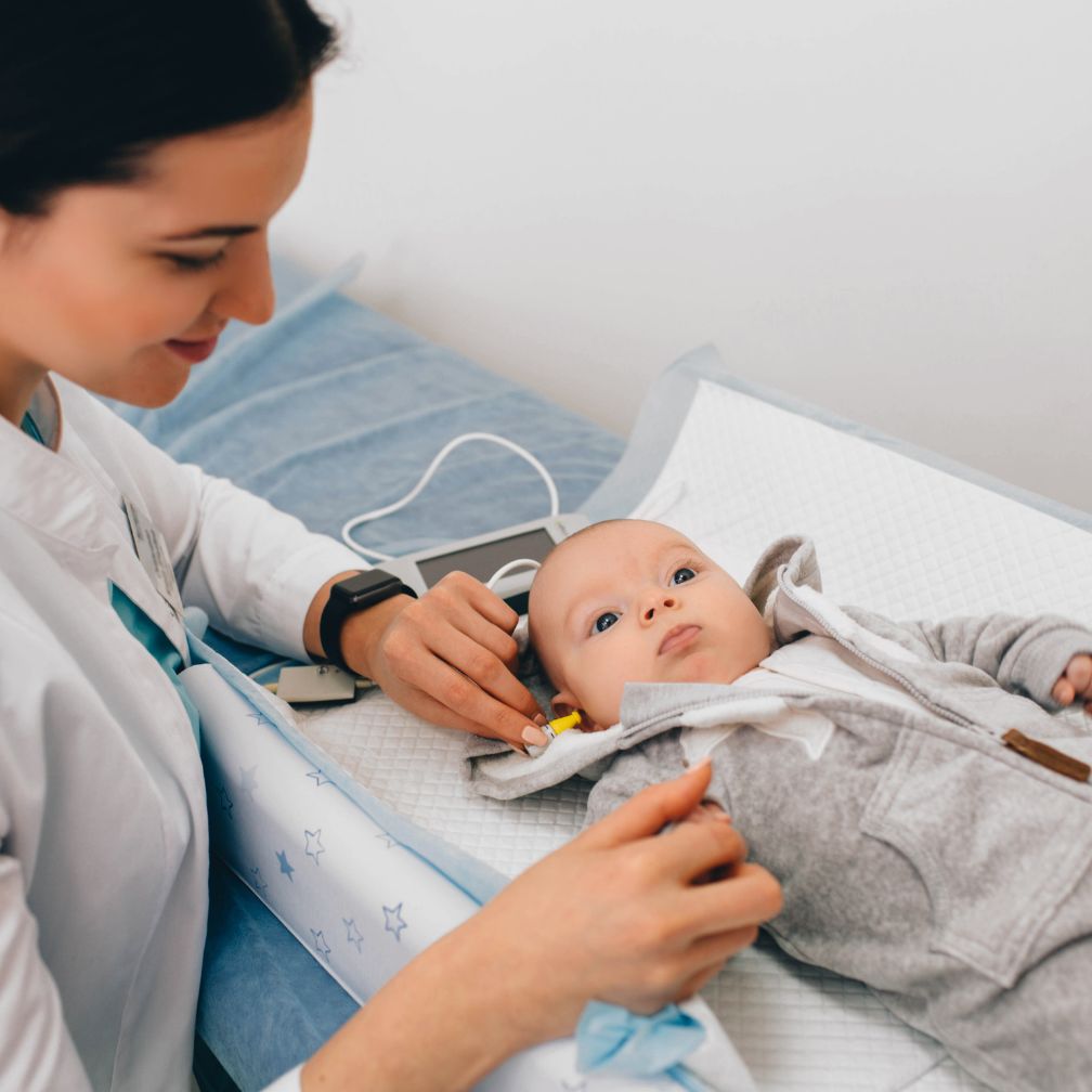 A healthcare professional performs a hearing test on a baby lying on an examination table, using small earphones and monitoring equipment.