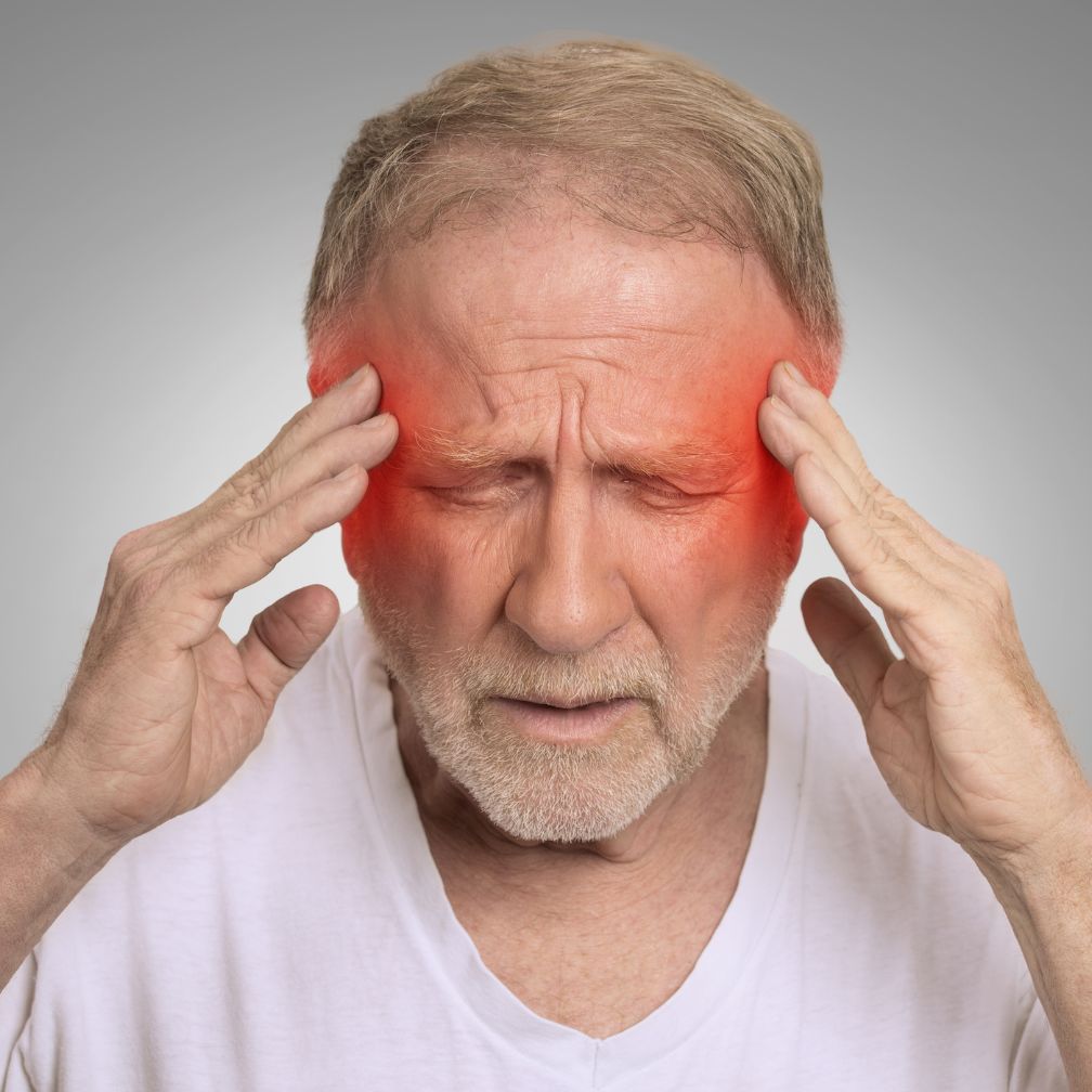 An older man with a beard holds his head, eyes closed, with red coloring on his temples, indicating pain or a headache.