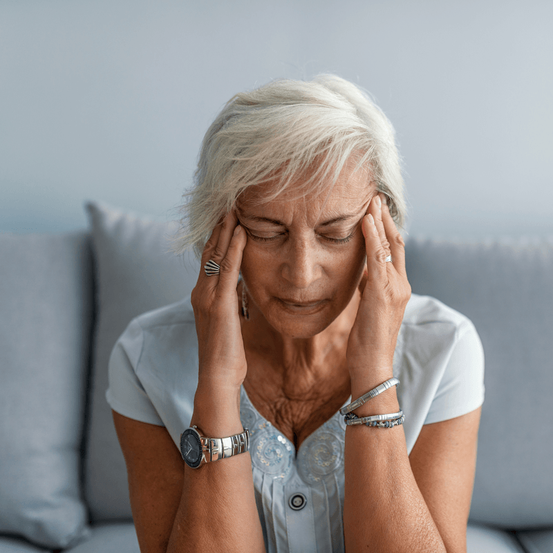 An older woman with short gray hair sits on a couch, eyes closed, pressing her temples with her fingers, appearing to have a headache or stress. She wears a light blue blouse, a watch, and several rings and bracelets.