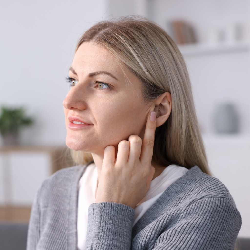 A woman with blonde hair touches her ear while looking to the side. She is wearing a gray sweater and is indoors, with a blurred background showing shelves and a plant.