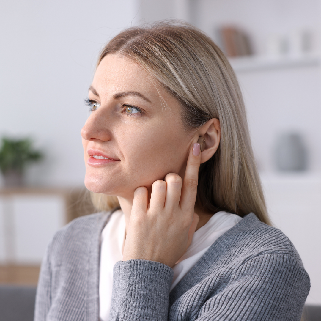 A woman with blonde hair touches her ear while looking to the side. She is wearing a gray sweater and is indoors, with a blurred background showing shelves and a plant.
