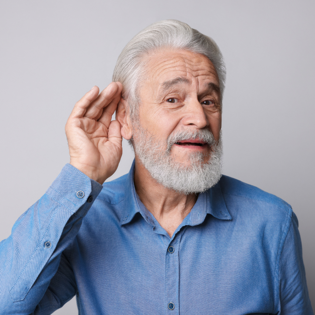 Older man with gray hair and beard wearing a blue shirt, holding his hand to his ear and leaning forward as if trying to hear something better, against a light gray background.